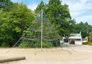 Einer Kletterspinne, also ein pyramidenförmiges Spielgerät aus Seilen zum Hochklettern für die Kinder. Im Hintergrund Bäume zu sehen.
