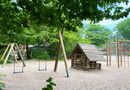 Eine Schaukel aus Holz, rechts daneben ein Holzspielhaus und ein Klettergerüst aus Holz auf Sandboden.