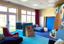 Ein Raum mit blauem Teppich und heller Fensterfront, in welchem verscheidene blaue Sofas verteilt stehen, ein Bücherregal, Kissen. An der rechten Seite erkennt man außerdem eine Hochebene aus Holz, mit verschiedenen Winkeln und Räumen in denen sich die Kinder aufhalten können.